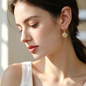 May include: A gold hoop earring featuring a small, textured seashell charm. The earring is crafted from polished gold metal, with the charm hanging from a circular hoop that secures through the earlobe, showcasing detailed ridges.