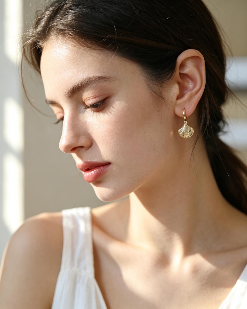 May include: A gold hoop earring featuring a small, textured seashell charm. The earring is crafted from polished gold metal, with the charm hanging from a circular hoop that secures through the earlobe, showcasing detailed ridges.