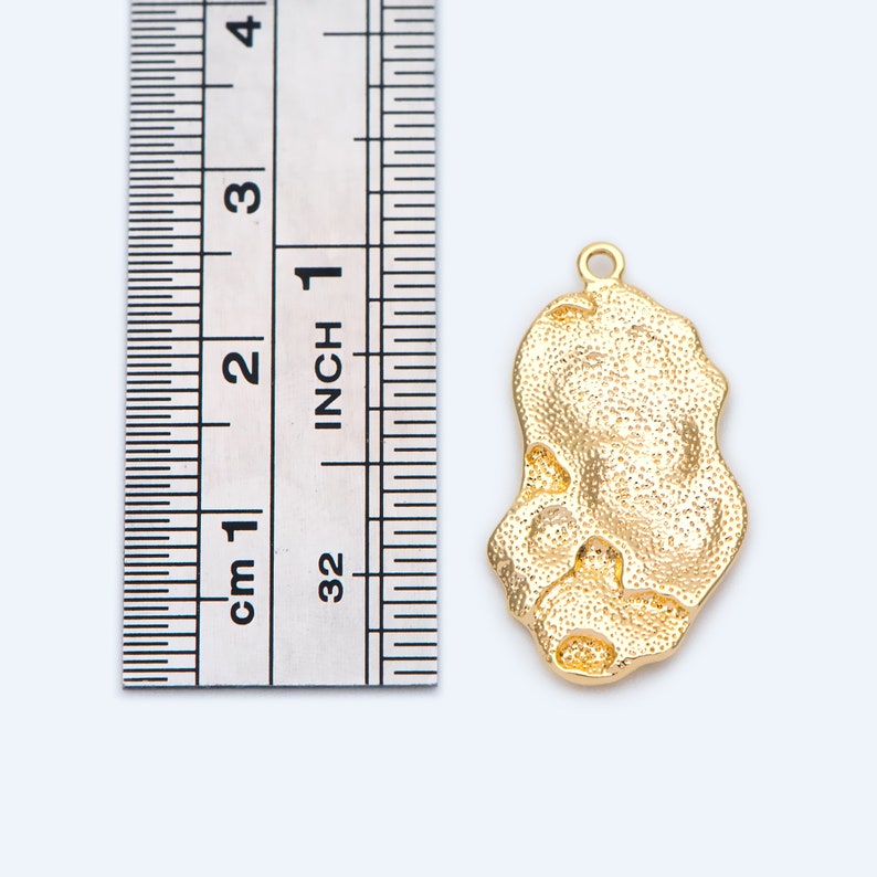 May include: Gold-toned metal pendant charm with a textured, irregular shape, measuring approximately 2.5 cm in length. The charm is shown next to a ruler for size reference.