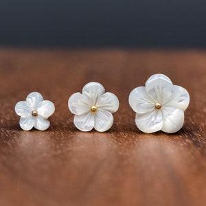 Peut inclure: Trois boucles d'oreilles en nacre blanche en forme de fleur avec des centres dorés. Les boucles d'oreilles sont de tailles différentes, la plus grande mesurant environ 2,5 cm de diamètre.