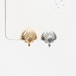 May include: Two seashell-shaped stud earrings, one gold-toned and one silver-toned, are displayed on a white card. The earrings have a detailed, fan-like design and a small loop at the bottom. The background is plain white.