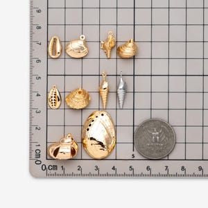 May include: A collection of gold and silver-toned metal shell charms arranged on a grid mat. The charms include various spiral, conch, and bivalve shapes. A US quarter coin is placed next to the charms for scale on the centimeter grid.