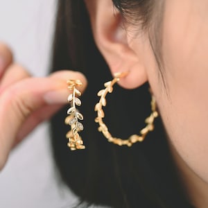 May include: Gold-colored hoop earrings with a leaf design. One earring is held up, showing the delicate details of the leaves and small, clear stones. The other earring is worn on the ear, showcasing the hoop shape.