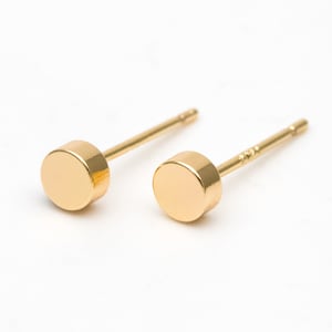 May include: A pair of gold-toned stud earrings with a round, flat design. The earrings have a simple, minimalist style.