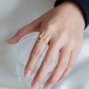 May include: A gold snake ring with a textured finish. The ring is worn on the index finger of a hand.