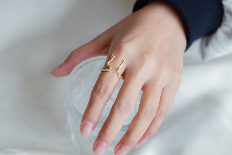 May include: A gold snake ring with a textured finish. The ring is worn on the index finger of a hand.