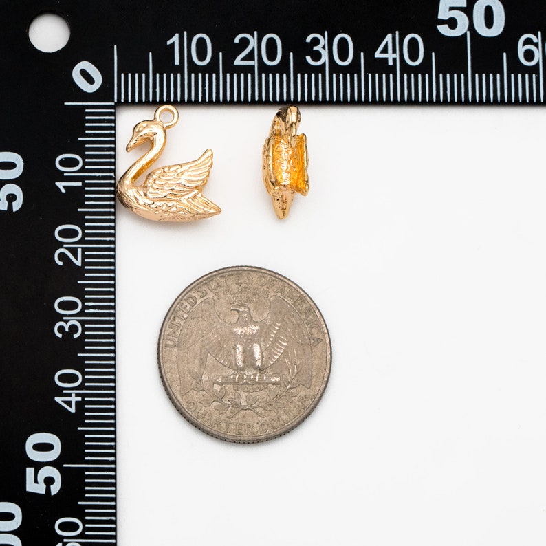 May include: Two gold-tone metal charms, one shaped like a swan and the other a more abstract design, are shown next to a US quarter for size comparison.