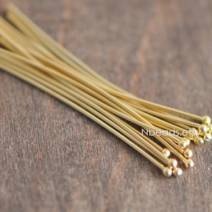 May include: A bundle of gold-colored head pins with rounded ends. The pins are arranged in a fan shape on a wooden surface.