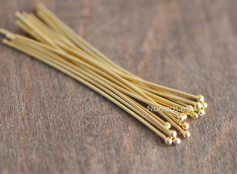 May include: A bundle of gold-colored head pins with rounded ends. The pins are arranged in a fan shape on a wooden surface.