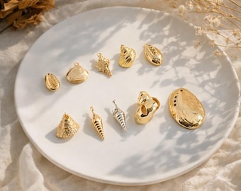 Op de afbeelding: Een verzameling van tien metalen bedels in de vorm van diverse schelpen, waaronder conch-, spiraal- en mosselontwerpen. Negen bedels hebben een gepolijste goudkleurige afwerking, terwijl één slanke spiraalbedel een zilverkleurige afwerking heeft.