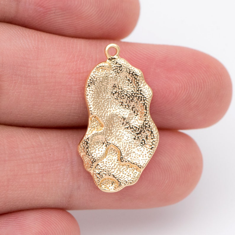 May include: Gold-toned metal pendant charm with an irregular, organic shape. The pendant has a textured surface and a loop at the top for attaching to a chain or cord.