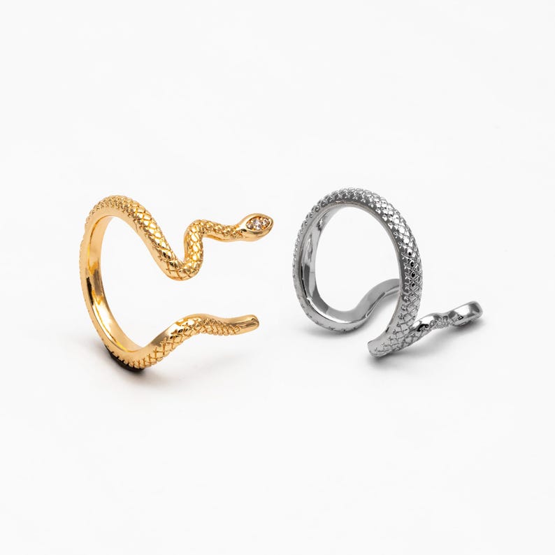 May include: Two snake-shaped rings, one gold and one silver. Both rings feature a textured snake design and a small clear stone on the head.