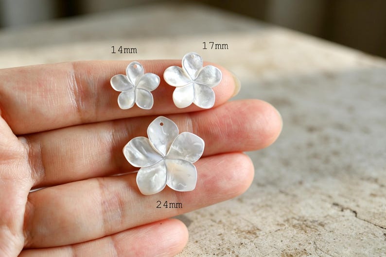May include: Three white, iridescent, flower-shaped charms with five petals each. The charms are labeled with their respective sizes: 14mm, 17mm, and 24mm. Each piece features a smooth, pearlescent finish and a small hole for attachment purposes.