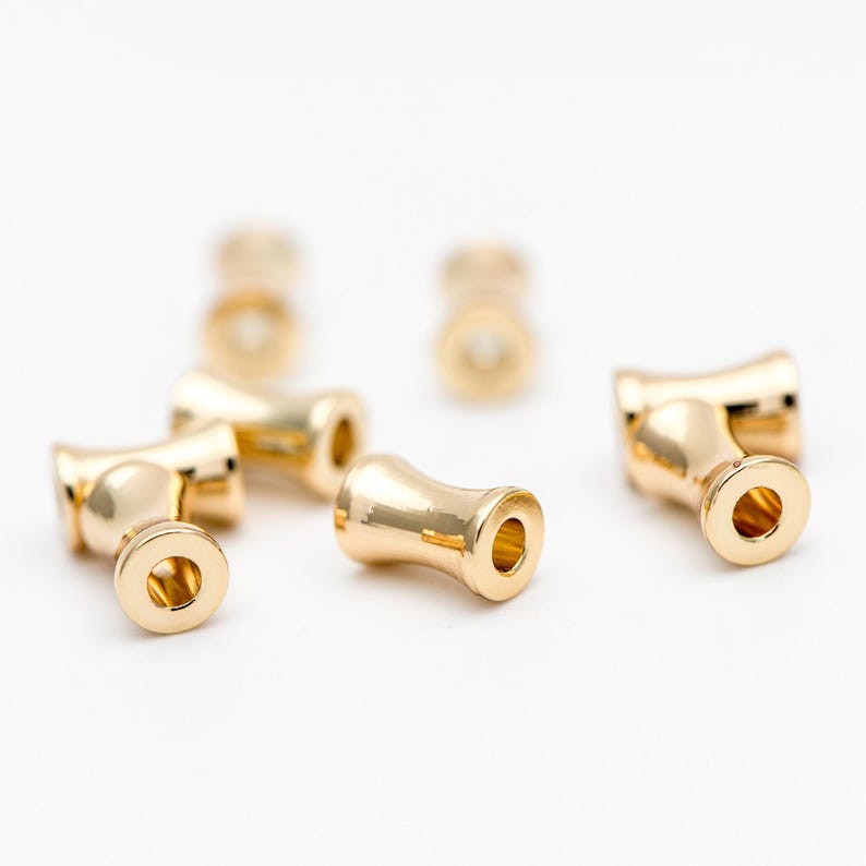 May include: Gold-toned metal beads with a cylindrical shape and a small hole in the center. The beads have a smooth, polished finish.