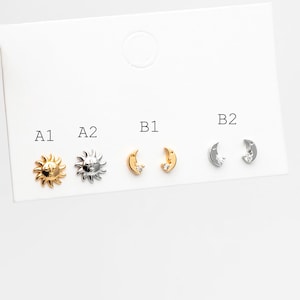 May include: A selection of stud earrings in gold and silver tones. Designs include sun and moon shapes, some with small crystal accents. The earrings are displayed on a white card.