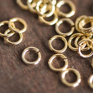 May include: A close-up of a pile of gold jump rings. The rings are small and round, and they are all made of the same metal. They are scattered on a wooden surface.