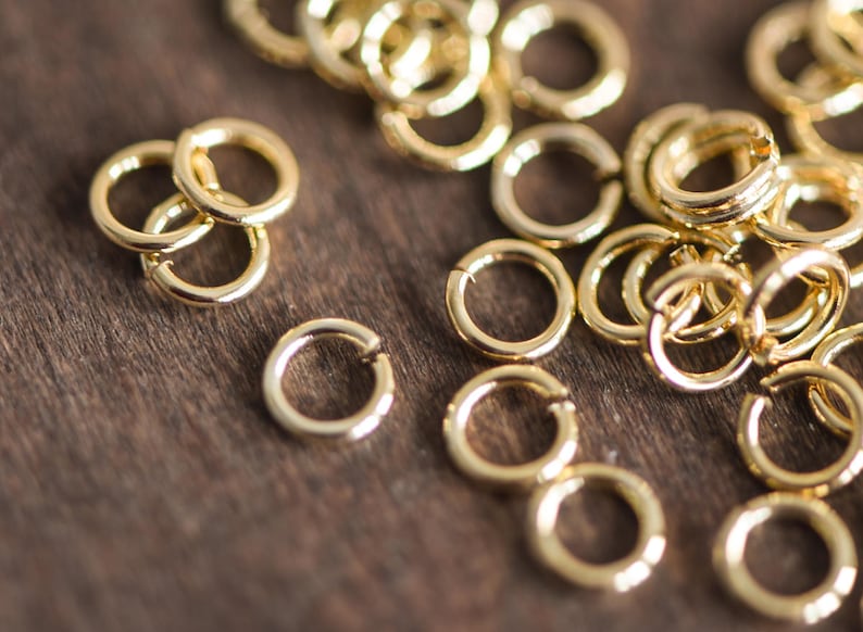 May include: A close-up of a pile of gold jump rings. The rings are small and round, and they are all made of the same metal. They are scattered on a wooden surface.