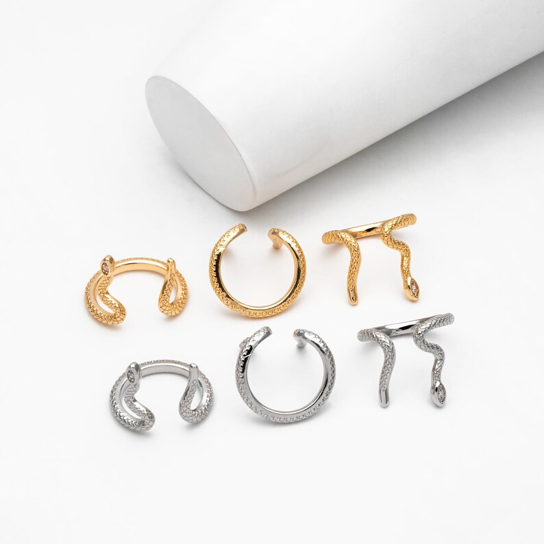 May include: Six gold and silver snake-shaped rings. The rings are open and have a textured surface. Some of the rings have a snake head with a small stone on the top.