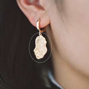 May include: Gold hoop earrings with a dangling irregular shaped charm. The charm is textured and has a slightly rough surface.