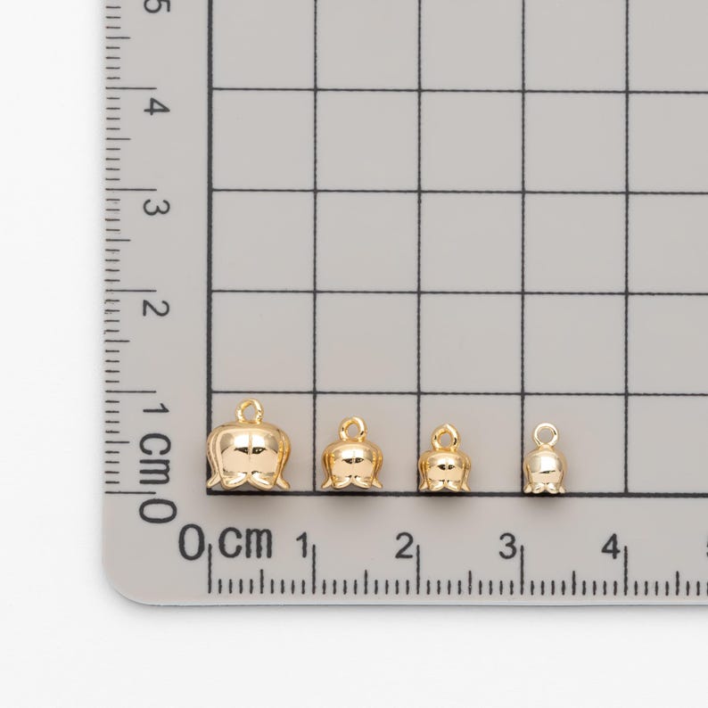 May include: Four gold-colored bell charms of varying sizes, measured in centimeters on a ruler.