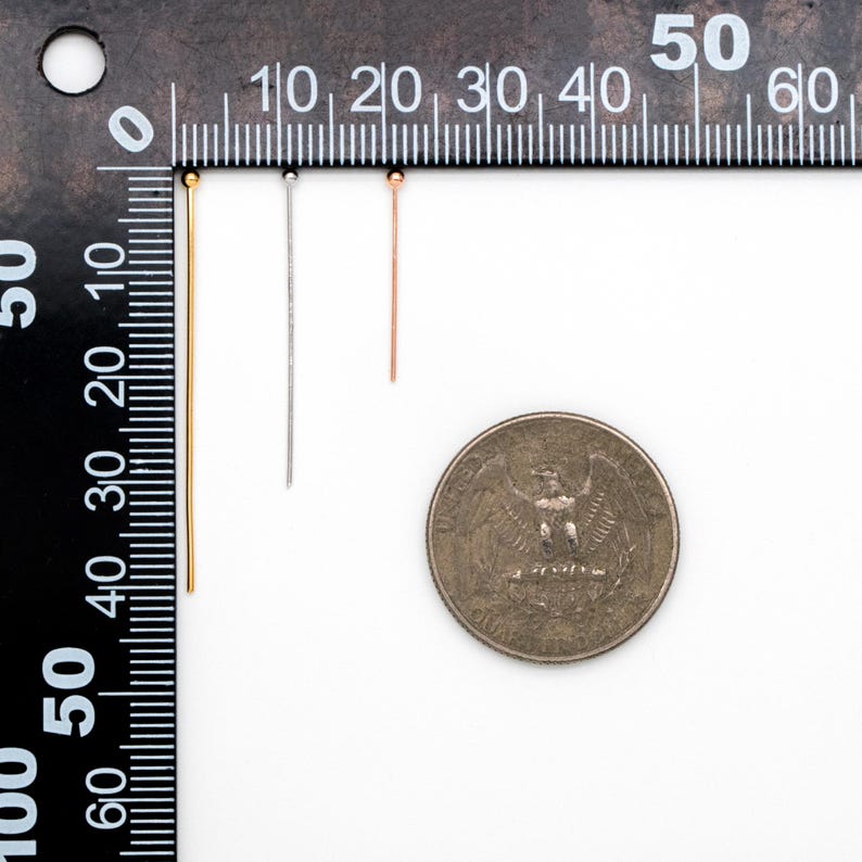 May include: Three thin metal headpins, one gold, one silver, and one rose gold, are lined up next to a ruler. The headpins are approximately 2 inches long. A US quarter is shown for scale.