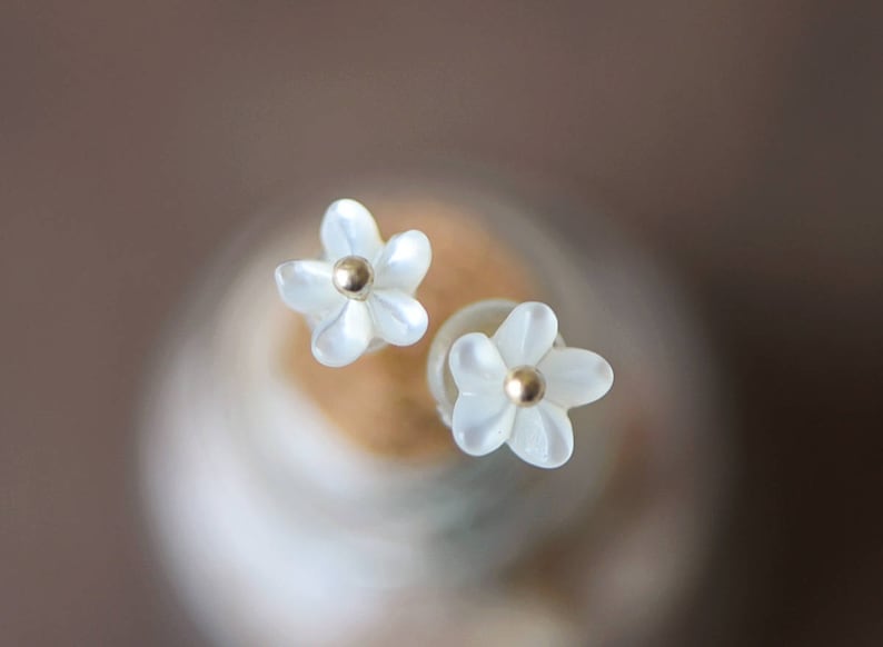 K&ouml;nnte beinhalten: Ein Paar wei&szlig;e, blumenf&ouml;rmige Ohrringe mit goldenen Zentren. Die Ohrringe sind klein und zart.