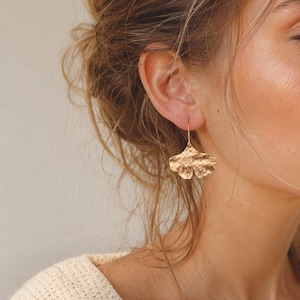 May include: Gold-colored leaf-shaped earrings. The earrings have a textured surface and a hook closure. The earrings are hanging from a person's ear. The earrings are a nature-inspired design.
