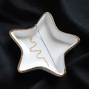 Può includere: Un piatto bianco a forma di stella con un bordo dorato, che contiene due gioielli. Uno è una sottile catena d'argento, l'altro è una catena d'oro con un design ondulato.