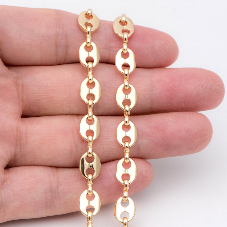 May include: Close-up of two strands of gold-colored chain. The chain links are oval-shaped with a central bar, creating a coffee bean or puffed link design. The chains are held against a light background.