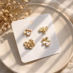 May include: A set of four gold-tone stud earrings displayed on a white card. The designs include a bee with a pear-shaped crystal, a bee with pavé crystals, a hexagonal honeycomb structure with small clear stones, and a flower with a crystal center.