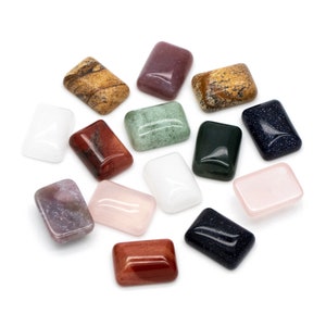 May include: A collection of fifteen polished, rectangular cabochon gemstones in various colors and textures, including white, pink, dark red, brown, green, and dark blue with glittery flecks, arranged in a scattered pattern on a white background.
