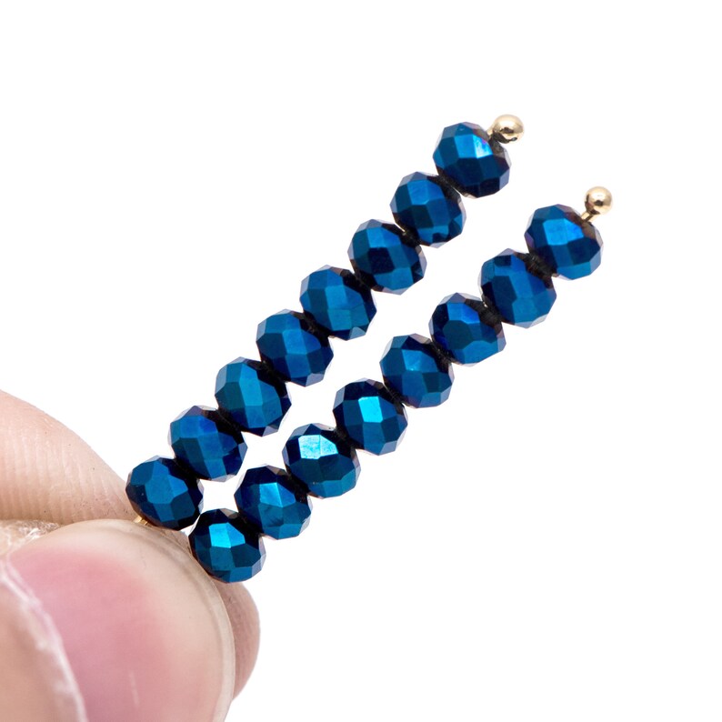 Pu&ograve; includere: Due fili di perline blu scuro sfaccettate, ciascuno con una piccola perla color oro all'estremit&agrave;. Le perline hanno una lucentezza metallica e sono infilate insieme. Lo sfondo &egrave; bianco.