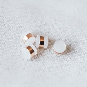 May include: Four white round beads with a rose gold band around the middle. The beads are on a white fabric background.