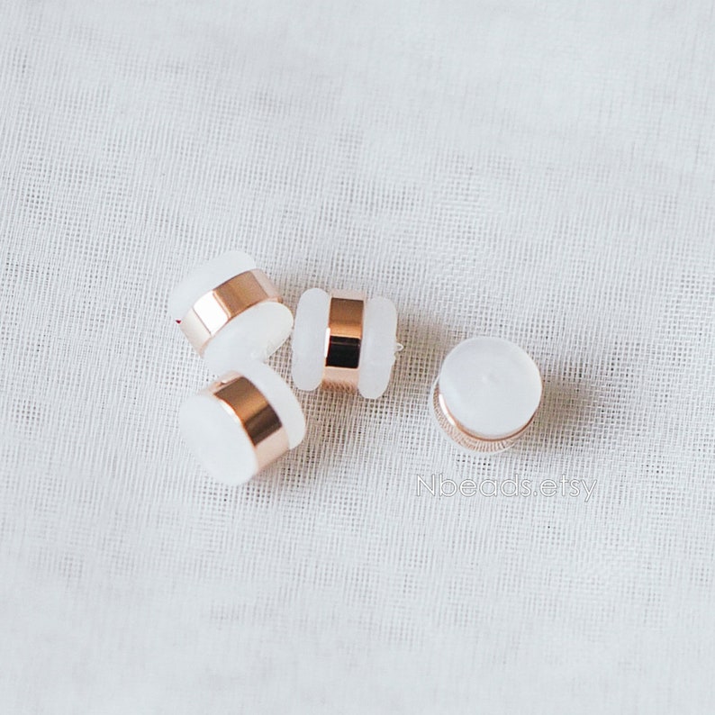 May include: Four white round beads with a rose gold band around the middle. The beads are on a white fabric background.