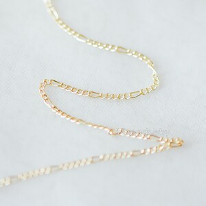 May include: Two gold chains, one on top of the other, are shown on a white surface. The chains are made of a delicate, figaro style link.
