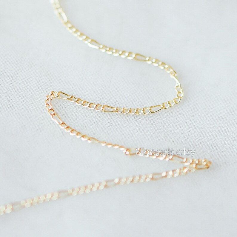 May include: Two gold chains, one on top of the other, are shown on a white surface. The chains are made of a delicate, figaro style link.