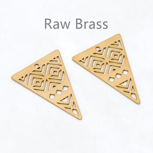 May include: Two raw brass triangle-shaped pendants with geometric cut-out designs. Each pendant has a small hole at the top for stringing. The text "Raw Brass" is above the pendants.
