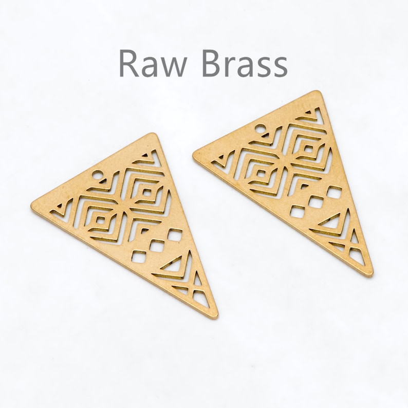 May include: Two raw brass triangle-shaped pendants with geometric cut-out designs. Each pendant has a small hole at the top for stringing. The text "Raw Brass" is above the pendants.