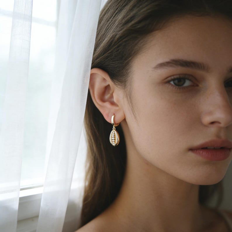 May include: A gold hoop earring featuring a small, dangling cowrie shell charm. The charm has a textured, ridged surface and is suspended from a polished gold hoop that secures through the earlobe, creating a delicate and refined jewelry piece.