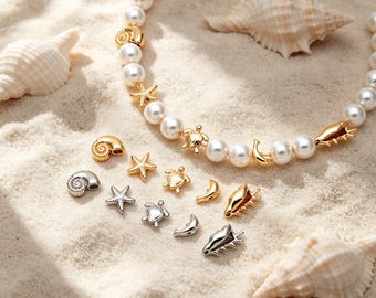 May include: A pearl necklace with gold seashell, starfish, and turtle charms. Additional charms are scattered on a sandy surface, along with seashells. The necklace and charms are in gold and silver tones.