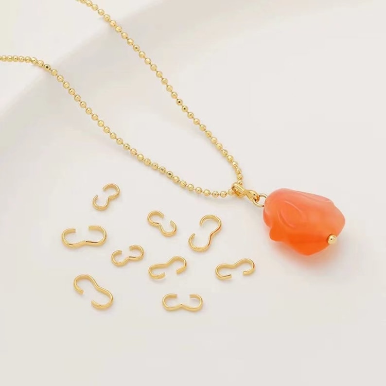 May include: A gold chain necklace with a peach-colored pendant shaped like a rose. The necklace is accompanied by several gold jump rings.