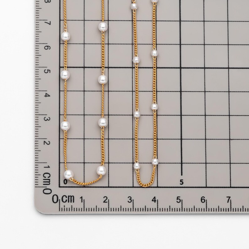 May include: Two gold chains with white pearl accents. The chains are laid out on a gridded surface with measurements in centimeters.