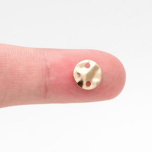 May include: A gold-coloured metal button with two holes. The button is round and has a slightly textured surface.