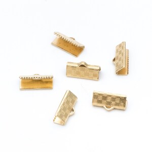 Puede incluir: Terminales de crimpado dorados dispersos sobre una superficie blanca. Los terminales tienen forma rectangular con patrones texturizados y bordes dentados. Algunos tienen un lazo para sujetarlos a otros componentes de joyer&iacute;a.