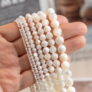 May include: A hand holds multiple strands of white mother-of-pearl beads. The beads vary in size, from small seed beads to larger, round ones. The beads have a smooth, pearlescent surface and a creamy white hue. The beads are suitable for jewellery making.