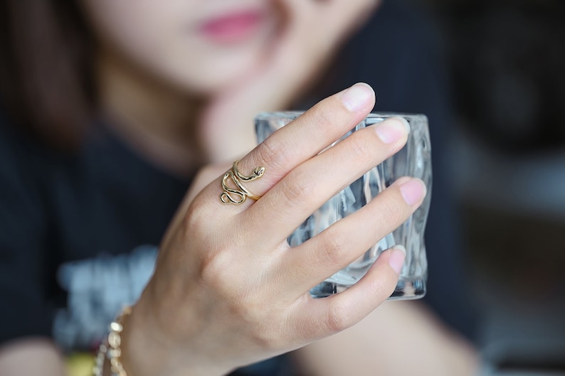 May include: A gold-coloured snake ring is wrapped around a finger. The ring has a coiled design with a snake head at the top. The ring is worn on a hand holding a clear glass.