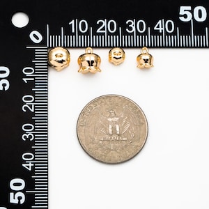 May include: Four gold-colored metal flower charms of varying sizes, measured against a ruler and a US quarter. The charms are in the shape of a bell with a rounded top and a small loop at the top.