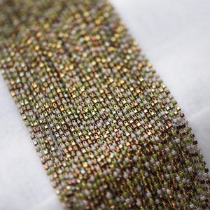 May include: A close-up of a chain made of small, square-shaped beads in shades of gold, green, and white. The beads are linked together to create a continuous chain.