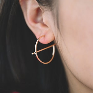 May include: Gold hoop earrings with a horizontal bar across the center. The earrings are worn in a person's ear.