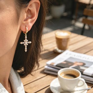Pu&ograve; includere: Orecchini a croce color oro con pietre preziose chiare e scintillanti. Gli orecchini sono appesi a un cerchio d'oro. L'immagine mostra un primo piano degli orecchini, con uno sfondo sfocato di un caff&egrave;.
